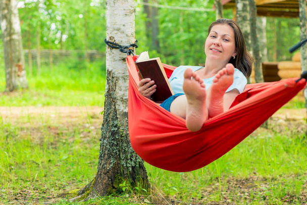 ευτυχισμένη γυναίκα 30 χρονών, διαβάζοντας ένα βιβλίο, κάθεται σε μια κόκκινη αιώρα - Φωτογραφία, εικόνα