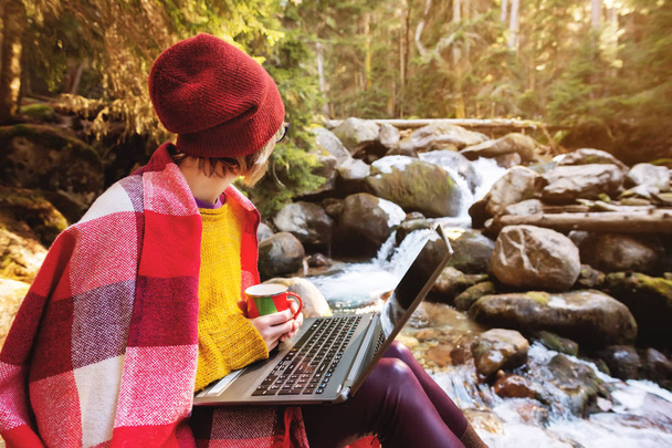 Hipster-Designerin mit kariertem Hut und gelbem Pullover und Brille mit Laptop auf dem Schoß und einer Tasse Kaffee in der Hand blickt auf einen Gebirgsfluss, der auf einem Felsen in einem - Foto, Bild