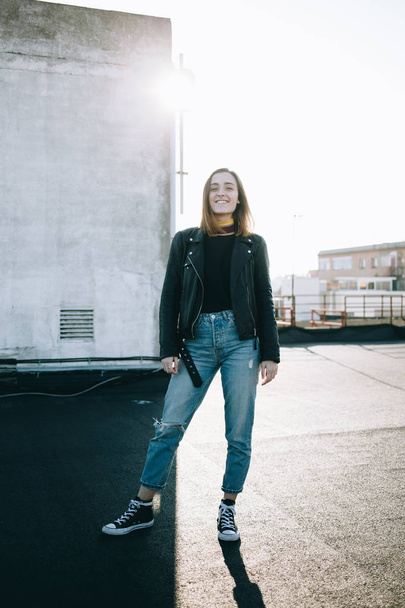 Wide shot of Empowered and independent attractive pretty woman or young teenager pose in fashionable stylish outfit, jeans and leather jacket on rooftop in sunset - Photo, image