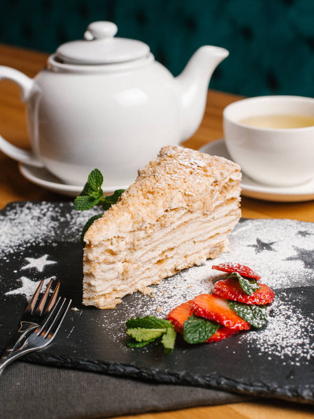 Lähikuva Napoleon kakku tarjoillaan kivi levy viipaloitu mansikka, minttu lehdet ja jauhettua sokeria, haarukat, teekannu ja kuppi teetä
 - Valokuva, kuva