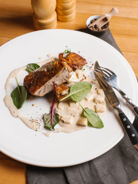  Жареная свинина с грибами и сливочным соусом подается со шпинатом и листьями свеклы на белой тарелке с салфеткой, вилкой, ножом, перцем и солью
 - Фото, изображение