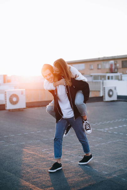 Young and attractive couple of millennials chill and relax on rooftop in sunset, laugh, smile and flirt, have fun doing piggy rides, fool around during date or romantic getaway, influencer or blogger - Foto, Imagem