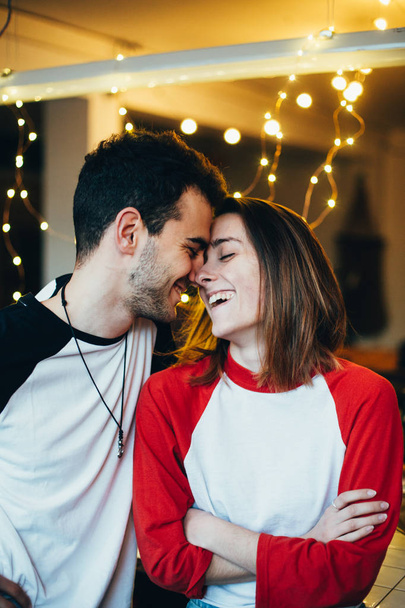 Happy and adorable, couple of cute young teenagers or millennials, laugh smile and cuddle inside apartment or studio with holiday lights, wear baseball shirts, excited about love and relationship - Foto, Imagen