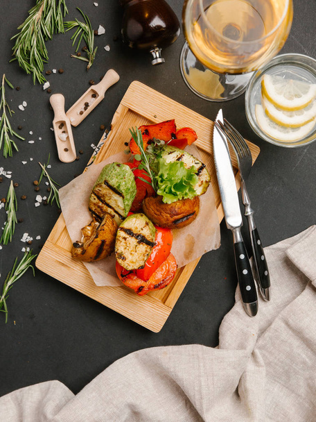  Grilled zucchini, potato, tomato and sweet red pepper served on parchment paper and wooden board with knife, fork, lemonade and wine - Photo, Image