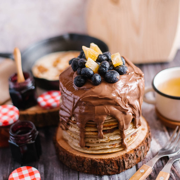 Nahaufnahme von Pfannkuchen mit Nutella, Blaubeeren und Orangenscheiben auf gesägtem Holz serviert mit einer Tasse Tee, Gabeln, Marmelade und Schneidebrett auf einem Holztisch - Foto, Bild