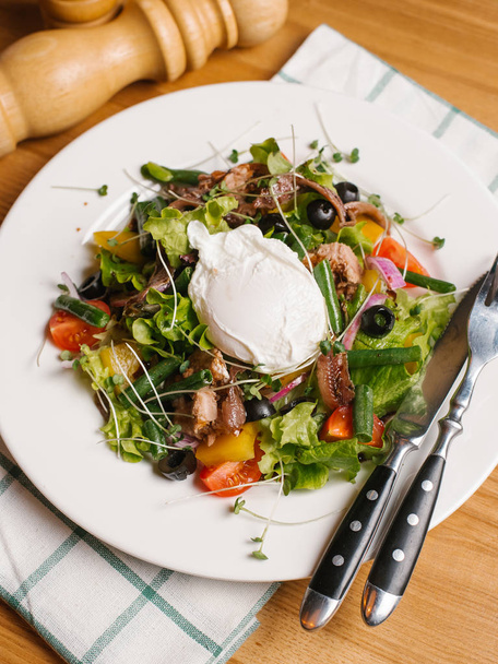 Salad with smoked eel, sweet peppers, cherry tomatoes, olives, lettuce, sweet onion, green beans and peas sprouts served on white plate with yoghurt, knife, fork, salt and pepper grinders - Photo, Image
