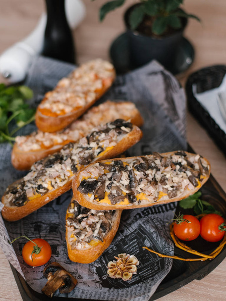 Hot sandwiches with mushrooms, ham, cheese, walnuts, mustard and ketchup served on wooden board with parsley and cherry tomatoes, fork and knife, vase, salt grinder and pepper grinder - Photo, Image