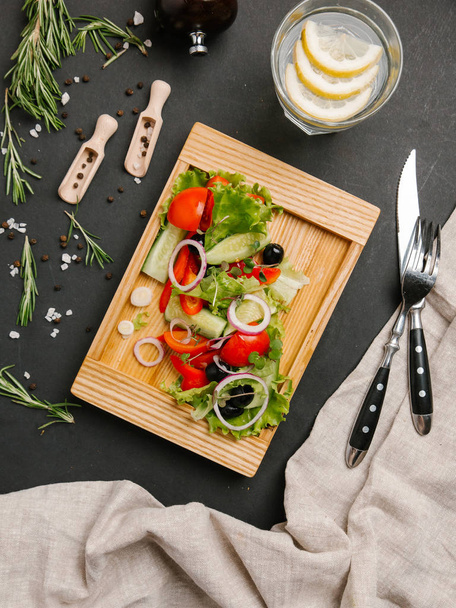 Fresh salad with sliced cucumber, tomato, sweet onion, olives and lettuce served on wooden dish with fork, knife and lemonade  - Photo, Image