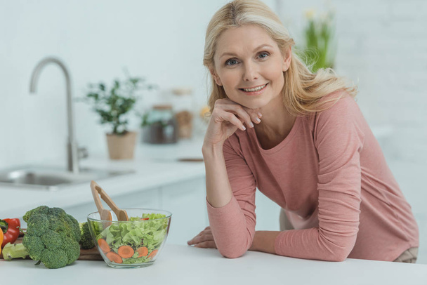 portrait of smiling mature woman with fresh salad on counter in kitchen - Фото, зображення