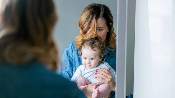Pelirroja joven madre de pie con su bebé encendido y mirando al espejo
 - Foto, imagen