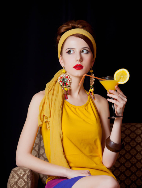 Young redhead girl in 70s yellow clothes style sitting in armchair with cocktail on black background - Photo, Image