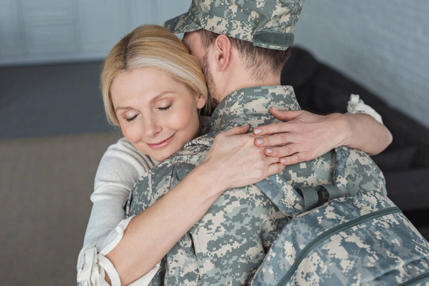 smiling mother hugging grown son in military uniform at home - Fotó, kép