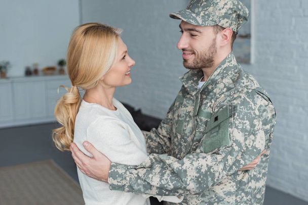 vista lateral de homem sorridente em uniforme militar e mãe abraçando uns aos outros em casa
 - Foto, Imagem
