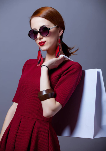 όμορφη νεαρή γυναίκα με κόκκινο φόρεμα και γυαλιά ηλίου που κατέχουν μόνιμη τσάντα για ψώνια σε γκρίζο φόντο  - Φωτογραφία, εικόνα
