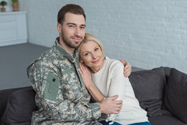 portrait of man in military uniform hugging smiling mother on sofa at home - Foto, Bild
