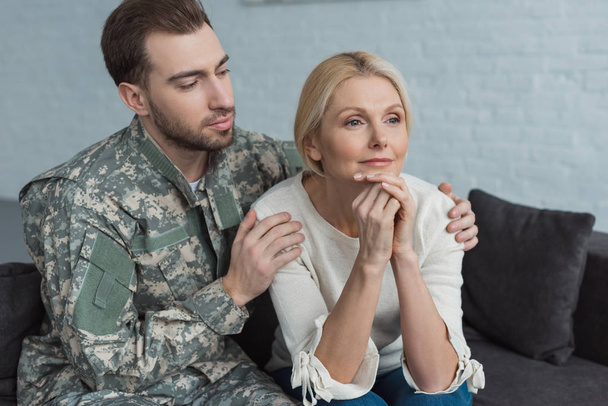 portrait grown son in military uniform hugging pensive mother on sofa at home - Photo, image