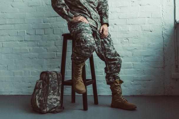 partial view of soldier in military uniform sitting on chair against white brick wall - Foto, immagini