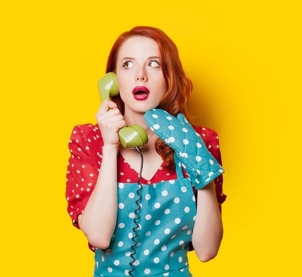 Redhead housewife with handset of dial phone on yellow background - Φωτογραφία, εικόνα