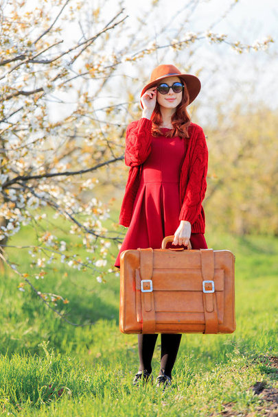 Redhead girl in red clothes and hat with suitcase in blossom cherry garden, full length - Foto, Imagem