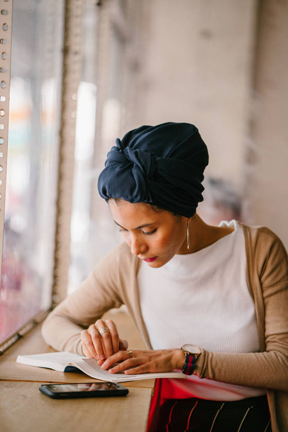 young, attractive Muslim woman (Arab, Malay, Asian) reads a book by the window of a cafe in the day. She is wearing a turban (headscarf, hijabi) and is elegantly dressed in earthy tones. - Foto, imagen