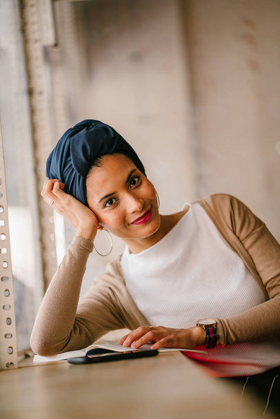 young, attractive Muslim woman (Arab, Malay, Asian) reads a book by the window of a cafe in the day. She is wearing a turban (headscarf, hijabi) and is elegantly dressed in earthy tones. - Photo, image