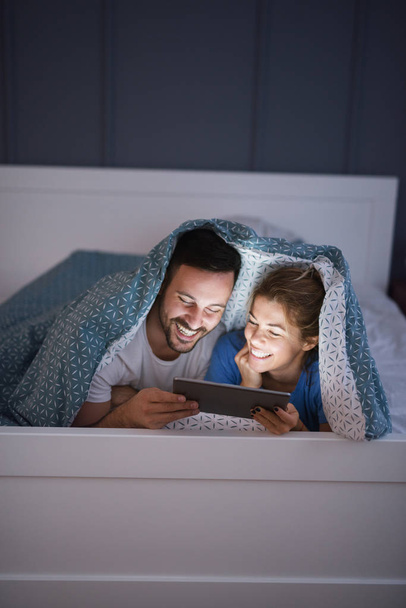 Szczęśliwa młoda miłość para leżąc w łóżku pod koc i oglądanie filmów za pomocą tabletu pc razem - Zdjęcie, obraz