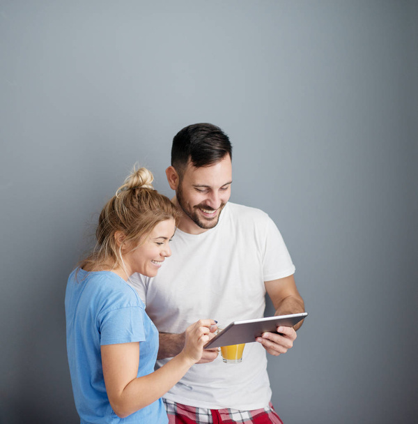glimlachend aantrekkelijke verliefde paar permanent samen in keuken in pyjama's en het lezen van nieuws via tablet pc voor het ontbijt  - Foto, afbeelding