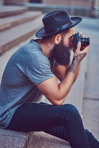 Красивый хипстер мужчина со стильной бородой и татуировкой на руках, одетый в повседневную одежду и шляпу держит камеру, сидит на ступеньках
. - Фото, изображение
