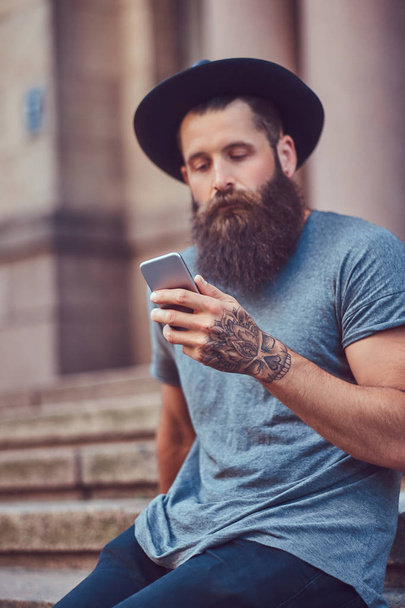 Hombre hipster guapo con una barba elegante y un tatuaje en su brazo vestido con ropa casual y sombrero usando teléfono inteligente mientras está sentado en los escalones
. - Foto, imagen