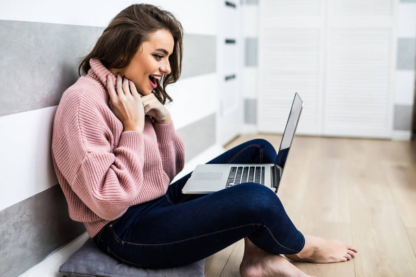 Gelukkig gillende jonge vrouw zittend op de vloer met laptopcomputer thuis - Foto, afbeelding