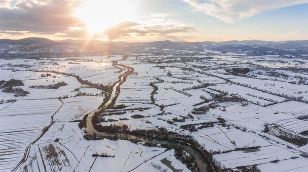 Vista aérea del río que fluye a través del campo cubierto de nieve al atardecer
. - Foto, imagen