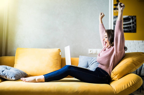 Pretty young woman using a laptop while raising hand and sitting on the couch - Fotografie, Obrázek