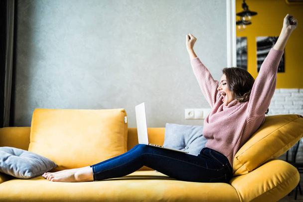 Νεαρή γυναίκα, σηκώνοντας τα χέρια της επάνω στον καναπέ με τον φορητό υπολογιστή στο σπίτι - Φωτογραφία, εικόνα