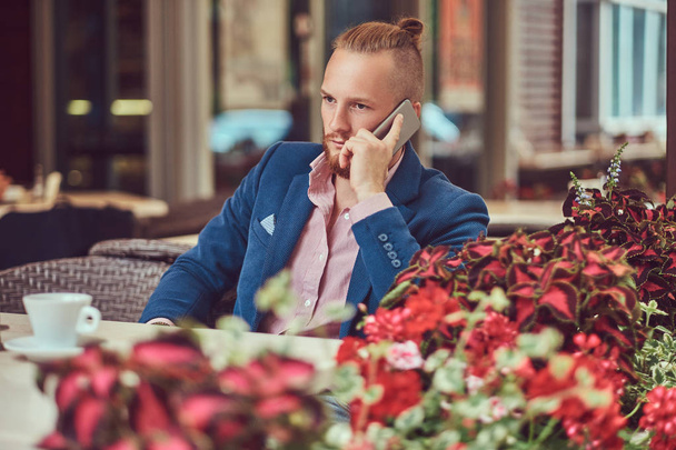 Handsome redhead businessman with a stylish haircut and beard dressed in a blue jacket and pink shirt, talking on the phone outside the cafeteria. - Foto, Imagen