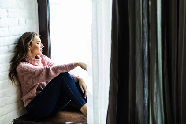 Χαμογελώντας η όμορφη γυναίκα στο πουλόβερ που κάθεται σε ένα windowsill στο σπίτι - Φωτογραφία, εικόνα