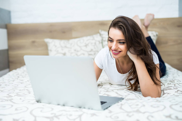 Mulher sorridente com as pernas levemente levantadas na frente de seu laptop deitado na cama
. - Foto, Imagem