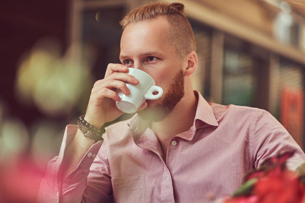 Hombre pelirrojo guapo con un corte de pelo elegante y barba en camisa rosa bebe café fuera de la cafetería
. - Foto, Imagen