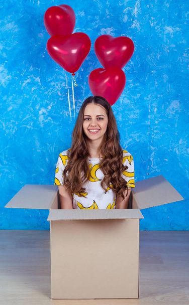 Wszystkiego najlepszego z okazji urodzin. Śmieszne, uśmiechnięta młoda dziewczyna siedzi w kartonie z darem i balony i patrząc na kamery w ubranie. - Zdjęcie, obraz
