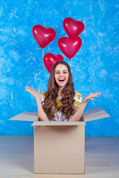 Happy Birthday. Funny smiling young girl sitting in a cardboard box with a gift and balloons and looking at the camera in casual clothes. - Foto, Imagem