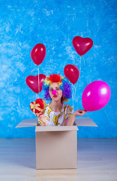 1 April Fools' Day concept. Funny clown sits in a cardboard box with helium balloons and looking in camera. Birthday gift. - Foto, immagini