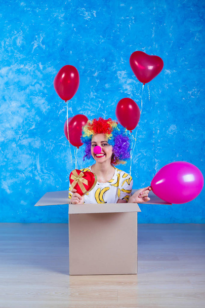 1 April Fools' Day concept. Funny clown sits in a cardboard box with helium balloons and looking in camera. Birthday gift. - Φωτογραφία, εικόνα