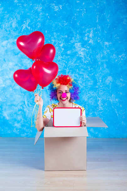 1 April Fools' Day concept. Funny clown sits in a cardboard box with helium balloons and looking in camera. Birthday gift. - Фото, изображение