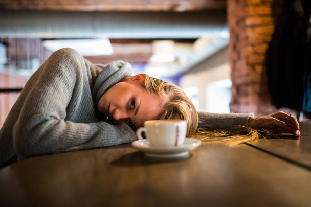 Beautiful young woman lies on hands, sits at wooden table in cafeteria, drinks coffee. Relax and rest concept - 写真・画像