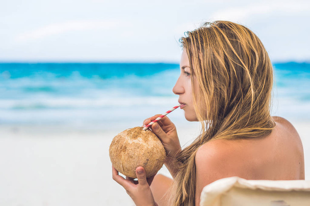 Young woman drinking coconut milk on Chaise-longue on beach. Dream escape with beauty girl. The Benefits of Coconut Water. - Foto, Bild
