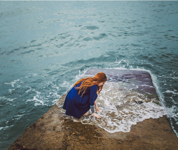 pretty red hair woman in blue dress sitting on sea shore - Fotoğraf, Görsel