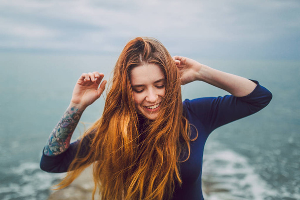 portrait of happy woman with long red hair in blue dress on sea shore at cloudy day - Photo, Image