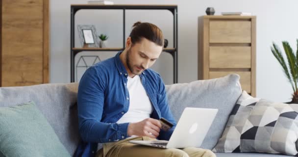 Young cheerful man in jeans shirt holding a credit card in a hand and buying online on the laptop computer on the sofa in the living room. Indoors - Πλάνα, βίντεο