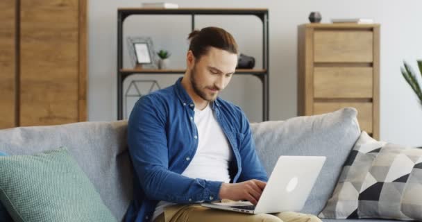 Young attractive man in jeans shirt sitting on the couch with a credit card in a hand and shopping online on the laptop computer in the cozy room. Inside - Footage, Video