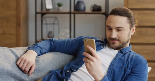 Portrait of the attractive man in jeans shirt resting on the sofa and texting and taping on the smartphone in the living room. Indoors - Кадры, видео