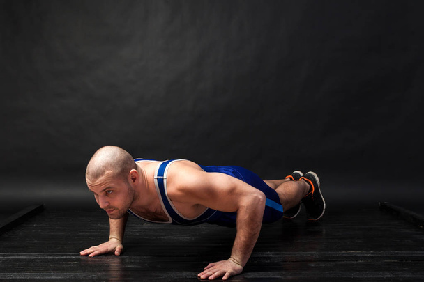 A young athletic man in blue wrestling tricky and blue shorts does push-ups on a black-haired isolated background in a photo studio - Foto, afbeelding